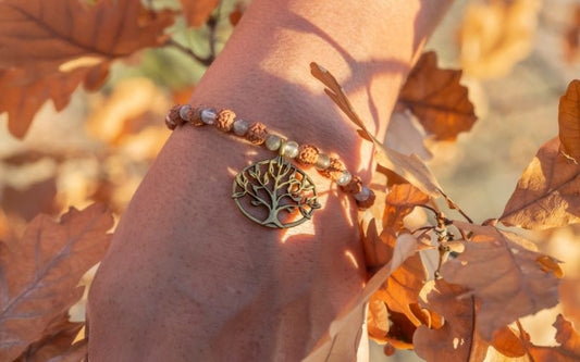 Femme qui glisse sa main entre des feuille morte marron, qui porte un bracelet avec un pendentif arbre de vie en cuivre. 