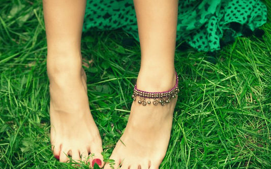 pieds et bas jambes d'une femme dans l'herbe verte qui porte un bracelet de cheville macramé avec des petites perles rondes et signes paece and love dorés