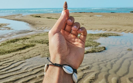 Main d'un homme avec un bracelet coquillage grain de café blanc, tenant un joli petit coquillage au bout des doigts et la mer, le sable en fond de la photo.