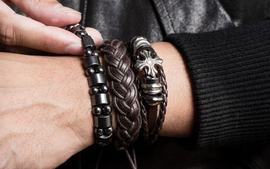 Poignet homme portant 3 bracelet dont 2 bracelets en cuir un tressé et un avec une genre de croix en acier inoxydable. Et un bracelet noir en perles