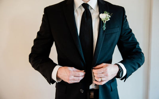 Homme en costume 2 pièces, avec cravate et fleur au col de la veste. Il porte un bracelet de montre noir très class pour aller avec le costume