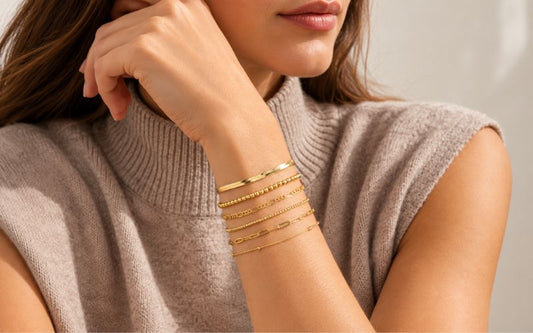 Femme qui pose, le bras plier vers son visage, elle porte un pull sans manches avec col roulé beige. ses cheveux son long, détachés et châtain avec des mèches claires. elle porte un lot de bracelets multirang