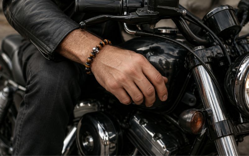 Poignet et main d'homme portant un bracelet œil de tigre, l'homme est sur une moto noire et habillé de noir. On ne voit qu'une partie de la moto et de l'homme.