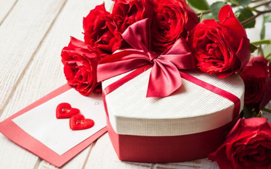 Bouquet de roses rouges avec boite cadeau rouge et blanche en forme de cœur et carte avec des cœurs posé sur une table blanche.