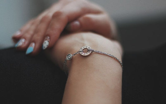 Femme qui croise les mains sur son ventre, on ne voit que ses mains et poignets, elle porte un bracelet chaine fine, avec un cercle gravé d'une inscription. 