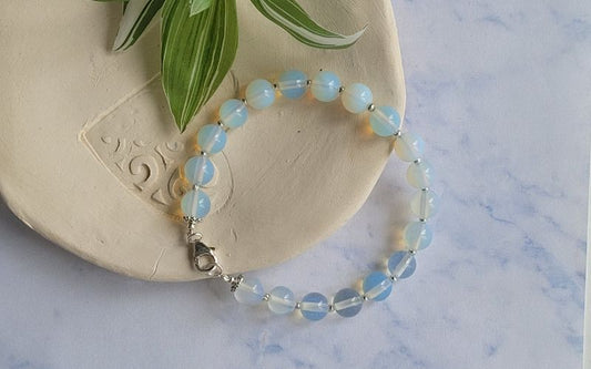 Bracelet en pierre de lune perles rondes, avec une accroche mousqueton présenté sur une pièce ovale d'argile beige et un bout de plante verte, on voit la table en marbre blanc gris en dessous