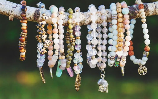 Plusieurs bracelet en pierres naturelles, enfilés sur un bâton, avec de la verdure en arrière plan flou. 